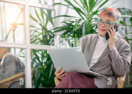 Senior businesswoman happily talking on phone with friend while sitting with laptop in business center with plants around. Wearing elegant blazer and Stock Photo