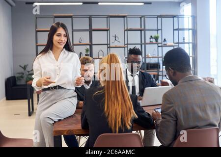 Informal meeting of business partners, caucasian and african people sit around table and discuss, while brunette with long female sitting on table and Stock Photo