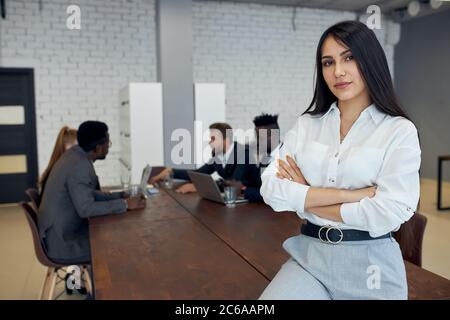 Caucasian beautiful woman wearing white formal shirt sit on table in office, confidently looking at camera