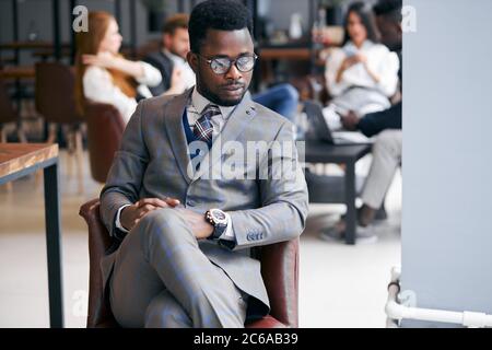 Portrait of handsome black man in eyeglasses wearing grey formal suit, elegant and serious businessman while his business partners have conversation b Stock Photo