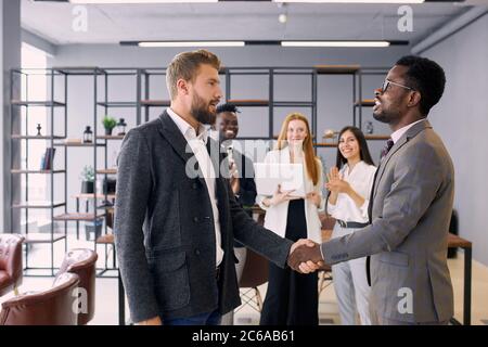 Two foreign executives in formal suits shaking hands in office while colleagues stand in background. Business people concept Stock Photo