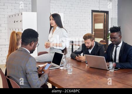 Informal meeting of business partners, caucasian and african people sit around table and discuss, while brunette with long female sitting on table. Is Stock Photo