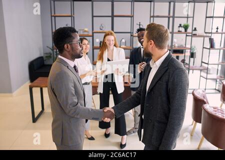 Two foreign executives in formal suits shaking hands in office while colleagues stand in background. Business people concept Stock Photo