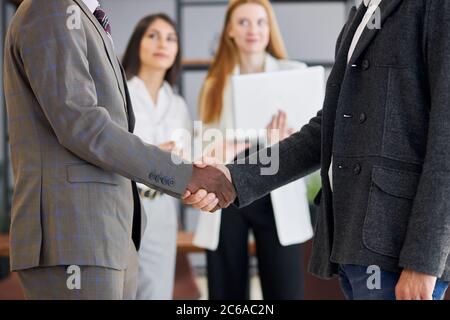 Two foreign executives in formal suits shaking hands in office while colleagues stand in background. Business people concept Stock Photo