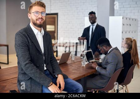 Attractive man confidently looking at camera while his business partners have discussion in office Stock Photo