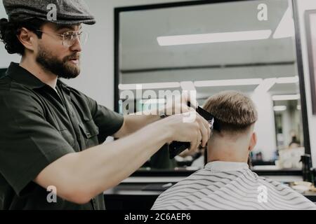 Young bearded man getting haircut by hairdresser while sitting in chair at barbershop Stock Photo