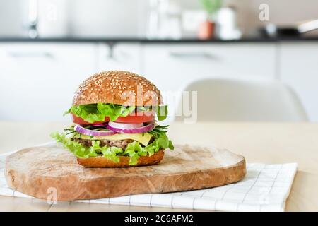 Beef burger with fresh salad, cheese and tomatoes on cutting board on the table in the bright kitchen. Fast food, unhealthy eating. Stock Photo