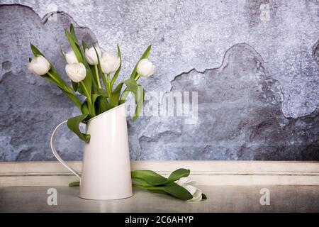 A tin jug filled with white tulips, over old plaster wall with grunge texture. Space for text Stock Photo