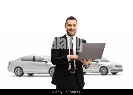 Man in a suit with a laptop computer smiling at the camera and posing in front of cars isolated on white background Stock Photo