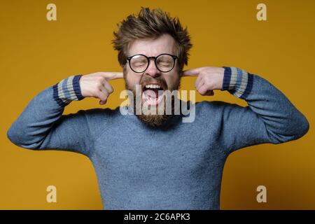 Man is stressed, noise annoys him, he covered ears with fingers, closed eyes and screams nervously. Stock Photo