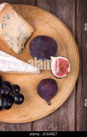 Old wooden chopping board,  with portions of cheese and fruit, over old wood table. Stock Photo