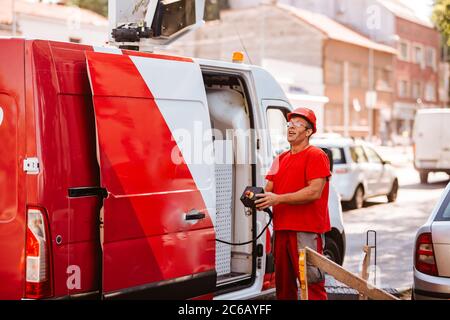 A middle-aged worker in a red uniform and helmet holds a control panel for lifting a crane and stands next to a van. Setting up traffic lights on the Stock Photo