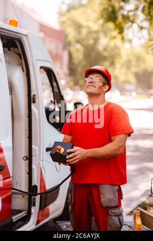 A middle-aged worker in a red uniform and helmet holds a control panel for lifting a crane and stands next to a van. Setting up traffic lights on the Stock Photo