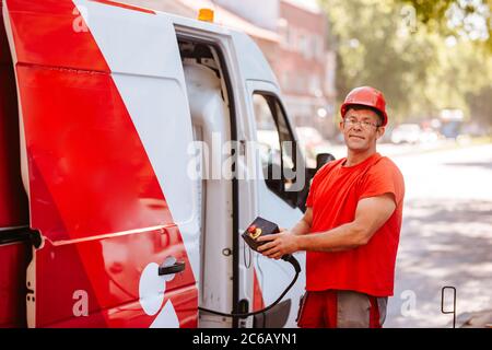 A middle-aged worker in a red uniform and helmet holds a control panel for lifting a crane and stands next to a van. Setting up traffic lights on the Stock Photo