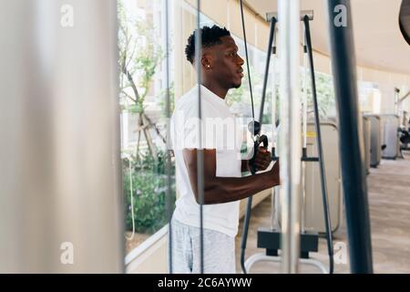 Muscular African American sportsman doing cable fly with exercise machine standing against window during training in gym. Stock Photo