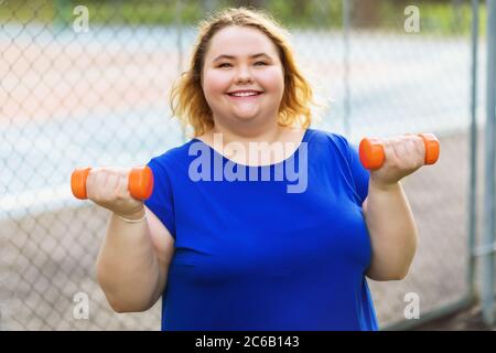 A young positive blonde of size plus performs exercises with dumbbells against the background of a sports ground. The concept of a healthy lifestyle Stock Photo