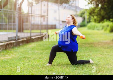 Young positive blonde size plus performs exercises with dumbbells in the park. The concept of a healthy lifestyle Stock Photo