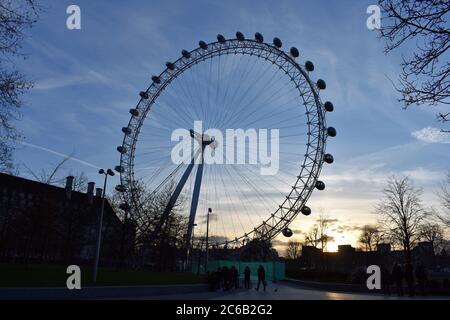 The London Eye at sunset along the South Bank of the River Thames.  A group of people can be seen by a green temporary fence below the wheel. Stock Photo