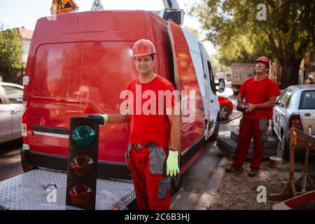 A middle-aged worker in a red uniform and helmet holds a control panel for lifting a crane, while in front of him stands a worker with a traffic light Stock Photo