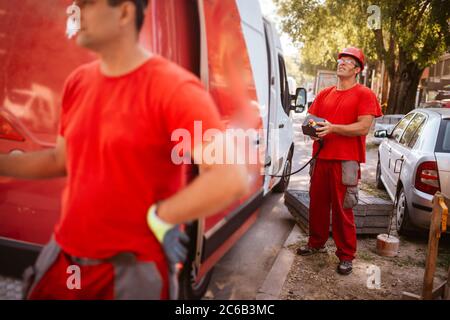 A middle-aged worker in a red uniform and helmet holds a control panel for lifting a crane, while in front of him stands a worker with a traffic light Stock Photo