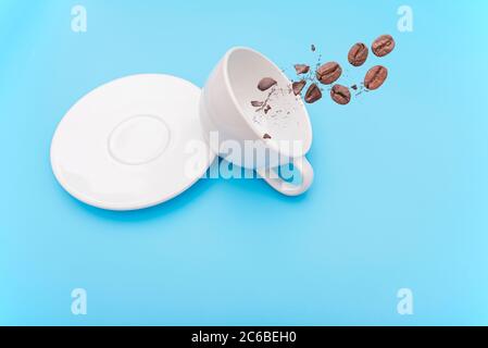 Overturned or knocked over white coffee cup, mug with bowl and coffee beans on blue background, concept picture Stock Photo