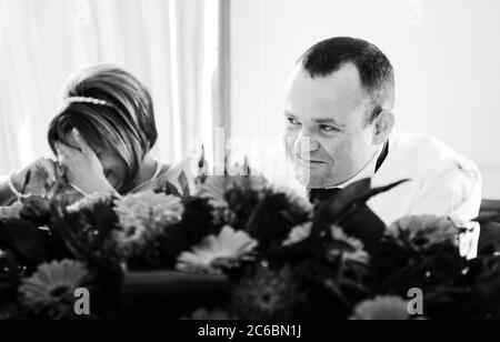 Bride facepalms while groom in his forties embarrassed laughs during wedding best man speech. Candid reportage real unstaged facepalm funny moment. Stock Photo