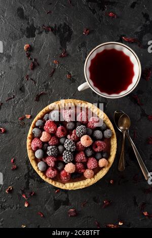Healthy dessert concept. Homemade cheesecake with frozen raspberries, blackberries and black currants served with hibiscus tea and flowers on black ba Stock Photo
