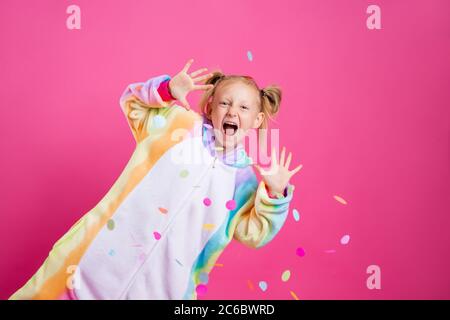 Happy little girl in kigurumi unicorn on a pink background rejoices in multi-colored confetti, space for text Stock Photo