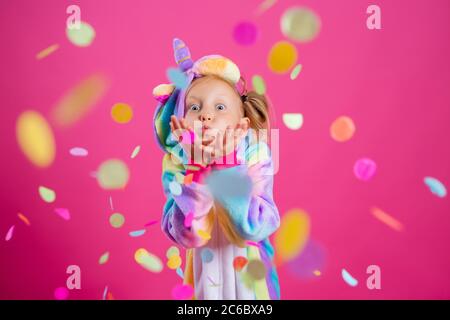 Happy little girl in kigurumi unicorn on a pink background rejoices in multi-colored confetti, space for text Stock Photo