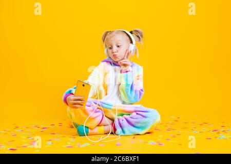 A happy little girl in a unicorn kigurumi sits on a yellow background taking a selfie. young blogger, space for text Stock Photo