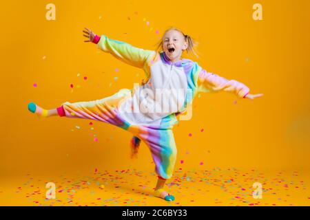 Happy little girl in kigurumi unicorn dancing on a yellow background among multicolored confetti Stock Photo