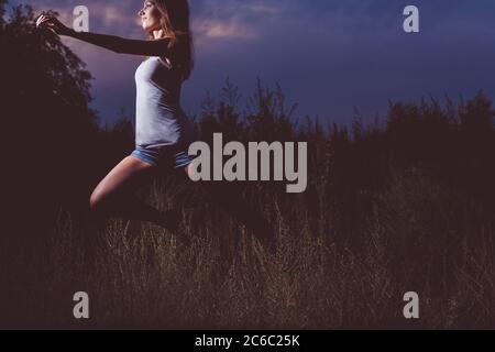 The sporty girl runs in the high grass at night. She dances or jumps full of young fever and energy. Stock Photo