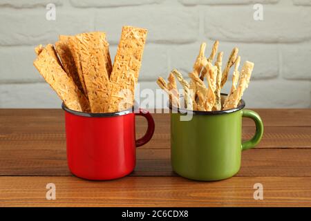 Grissini sticks and wheat bread with dried tomatoes, basil and cheese in enameled mugs standing on a brown wooden table. Closeup Stock Photo