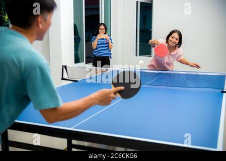 Couple fun playing table tennis or Ping pong indoor together leisure with competing in sports games at house. Father mother and daughter Asian family Stock Photo