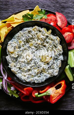 Spinach Artichoke leafy cheese Dip in a bowl with Tortilla Chips, cucumber, celery sticks, tomato slices and red bell pepper, vertical view from above Stock Photo