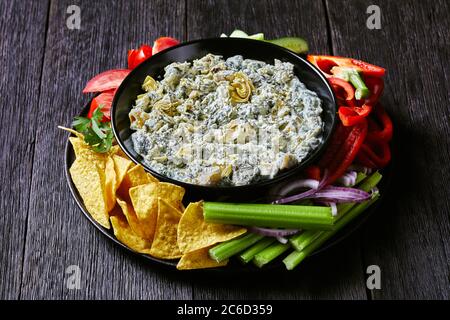 close-up of Spinach Artichoke leafy cheese Dip in a bowl with Tortilla Chips, cucumber, celery sticks, tomato slices and red bell pepper, landscape vi Stock Photo
