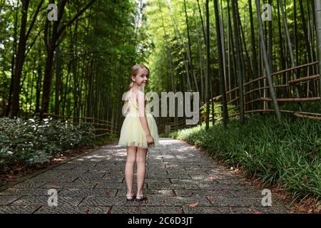 Cute little girl in yellow tutu dress standing in bamboo forest in China. Kid with blonde hair from behind outdoor Stock Photo