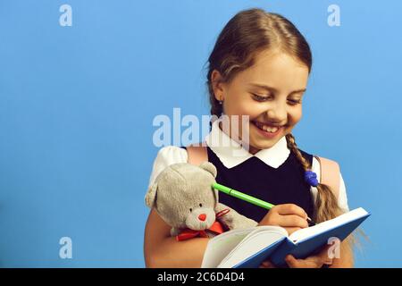 School girl with happy face isolated on blue background. Pupil in school uniform with braids and wide smile. Girl writes in big blue notebook with marker and holds teddy bear. Back to school concept Stock Photo