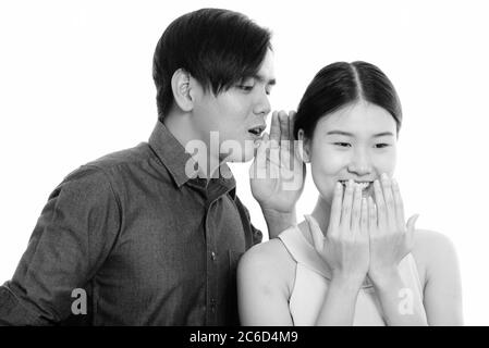 Studio shot of young Asian couple with man whispering to woman laughing Stock Photo