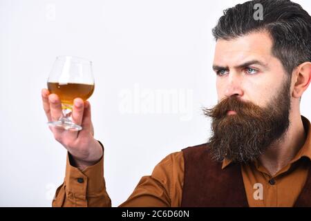 Barman in elegant uniform with serious face serves cognac. Waiter looks at whiskey or scotch in hand. Restaurant service concept. Man with beard holds glass with alcohol on white background, close up. Stock Photo