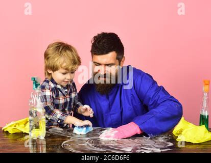 Man with smiling face and cute child on pink background. Guy with beard and mustache in rubber glove at table. Kid with father cleaning together with sponges. Parents little helper concept. Stock Photo