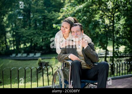 Happy bearded senior man in wheelchair with charming young girl supporting disabled grandparent on walking in park. Grandfather and granddaughter Stock Photo