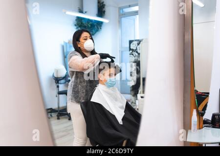 Reflection in the mirror of a hairdresser with a protective mask combing the hair of a boy with a protective mask sitting in a chair in a hairdressing Stock Photo