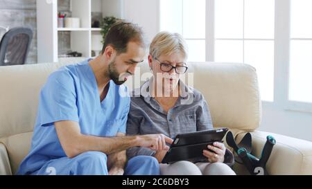 Male assistant teaching senior retired elderly old woman how to use her tablet computer in nursing home for her therapy Stock Photo