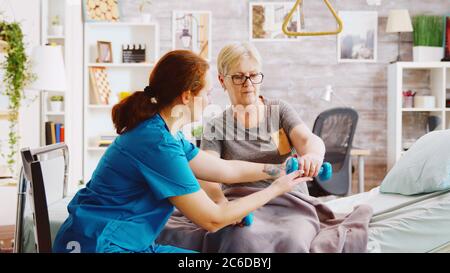 Revealing shot of female nurse helping an elderly woman to recover her muscles after an accident. She is lying in a hospital bed in a retirement home Stock Photo