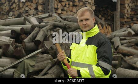 Portrait of lumberjack posing in green jacket with reflective stripes and holds small axe. Young blonde man woodcutter standing and looking at camera. Sawn logs, firewood background. Wood harvesting Stock Photo
