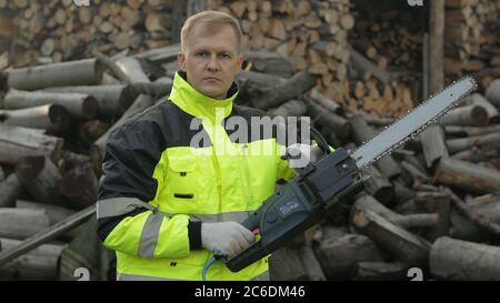 Portrait of lumberjack posing in green jacket with reflective stripes and holds electric chainsaw. Young man woodcutter standing and looking at camera. Sawn logs, firewood background. Wood harvesting Stock Photo