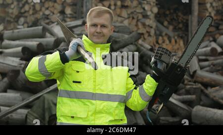 Portrait of lumberjack worker posing in green jacket with reflective stripes. Holds big axe and electric chainsaw in his hands. Man woodcutter. Sawn logs, firewood background. Wood harvesting Stock Photo