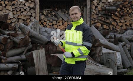 Lumberjack posing in green jacket with reflective stripes and holds small axe. Ready for work. Man woodcutter standing and looking at camera. Sawn logs, firewood background. Sawmill, wood harvesting Stock Photo