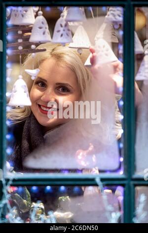A young blonde woman looks through a window pane that is frozen and adorned with Christmas decorations. The concept of Christmas time - image Stock Photo